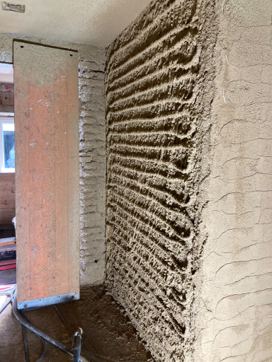 Strukturierter Lehmputz in horizontalen Rillen an der Wand, neben Holzrahmen – atmungsaktiv und texturiert. 